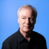 A man with gray hair is wearing a black shirt, looking slightly to the left against a plain blue background.