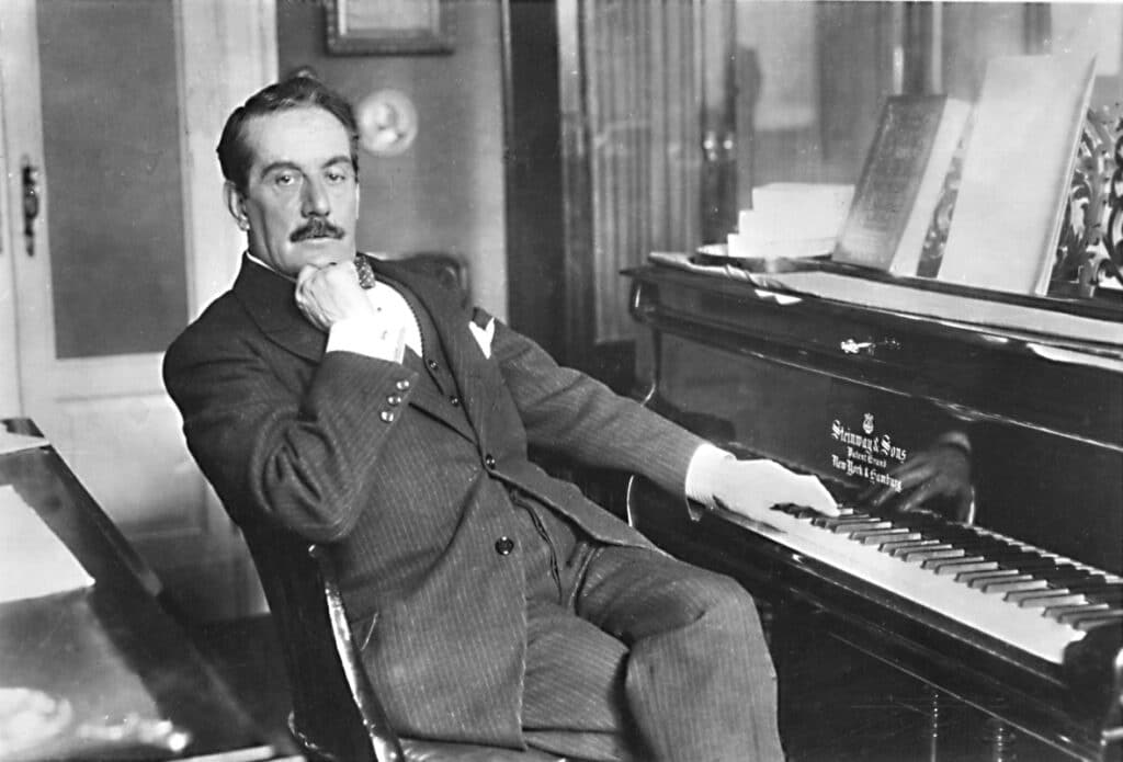 A black and white photo of a man with a moustache sitting sideways on a chair. His chin rests on his right hand while his left hand rests on the keys of a Steinway & Sons piano