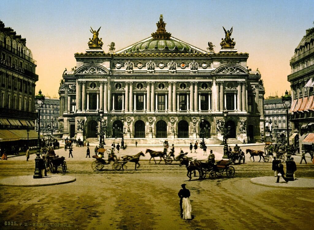 Front view of the Opéra Garnier, Paris, France displaying its ornate façade with golden statues on the rooftop with pedestrians and horse drawn carriages in front.