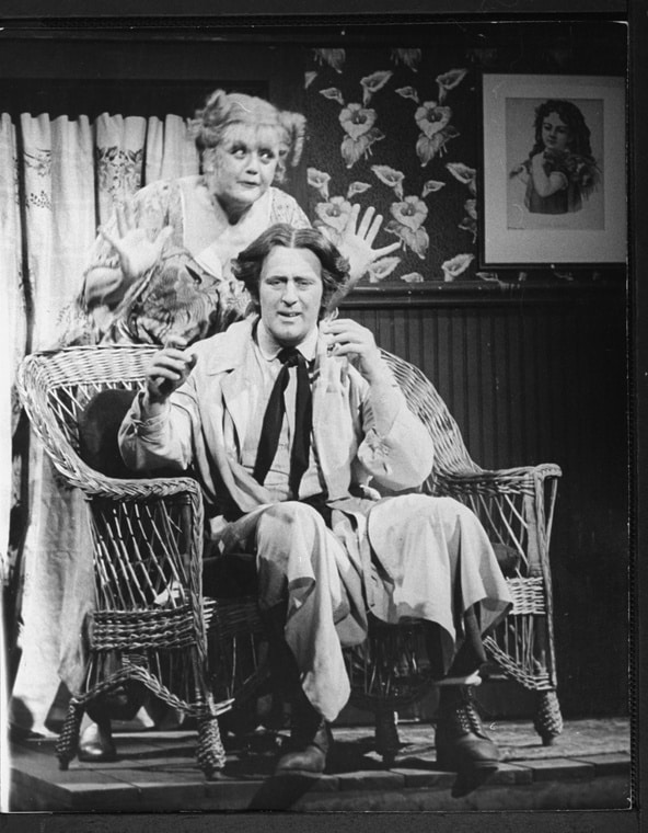 A man sitting on a wicker chair; behind him stands a woman making a playful expression. Both have their hands raised as if in mid-action. Black and white photo.