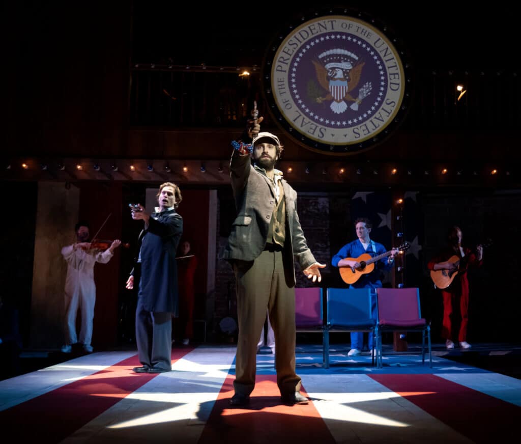 Several people stand on a floor painted like the American flag. The two nearest men are wearing old fashioned suits and aiming pistols, lit from above with star shaped spotlights. Five people wearing red, white, and blue jumpsuits stand in the back playing instruments. A large image of the Seal of the President of the United States hangs on the back wall.