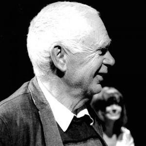 Profile of a middle-aged man with short white hair. Out of focus, a woman smiles behind him. Black and white photo.