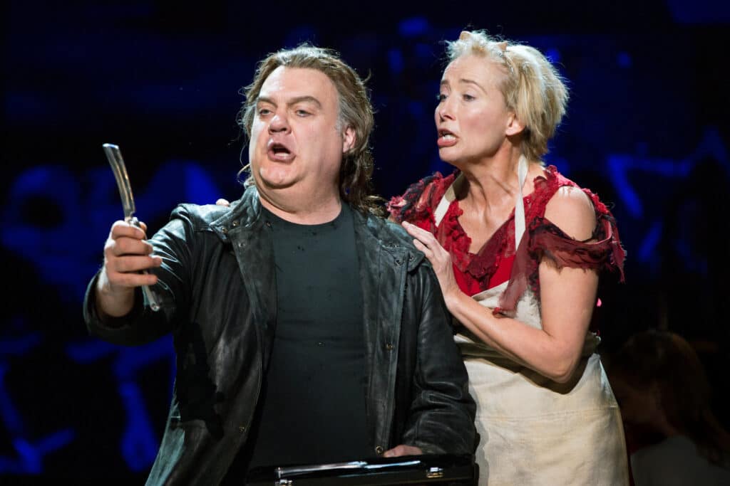 A man wearing black shirt and a black leather jacket holds up a straight razor. Behind him, a woman with short blond hair wearing a torn red dress and a white apron puts her hand on his shoulder. They are both staring at the razor, with their mouths open as if singing.