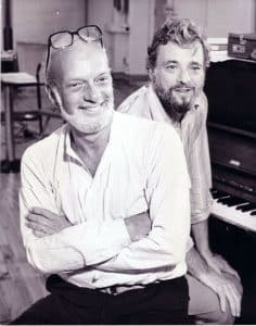 Two men sit on a piano bench smiling at the camera. The man in front is bald with a white beard and has his arms crossed and glasses on top of his head. The man behind, closer to the piano, has light hair and a beard and has his hands on his knees. Black and white photo.