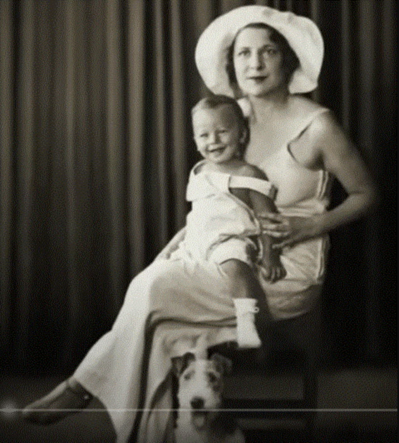 A woman in a long white sleeveless dress and a wide-brimmed hat sits in a chair. On her lap she holds a young smiling toddler who is also wearing white. A terrier sits on the floor in front of them. Black and white photo.