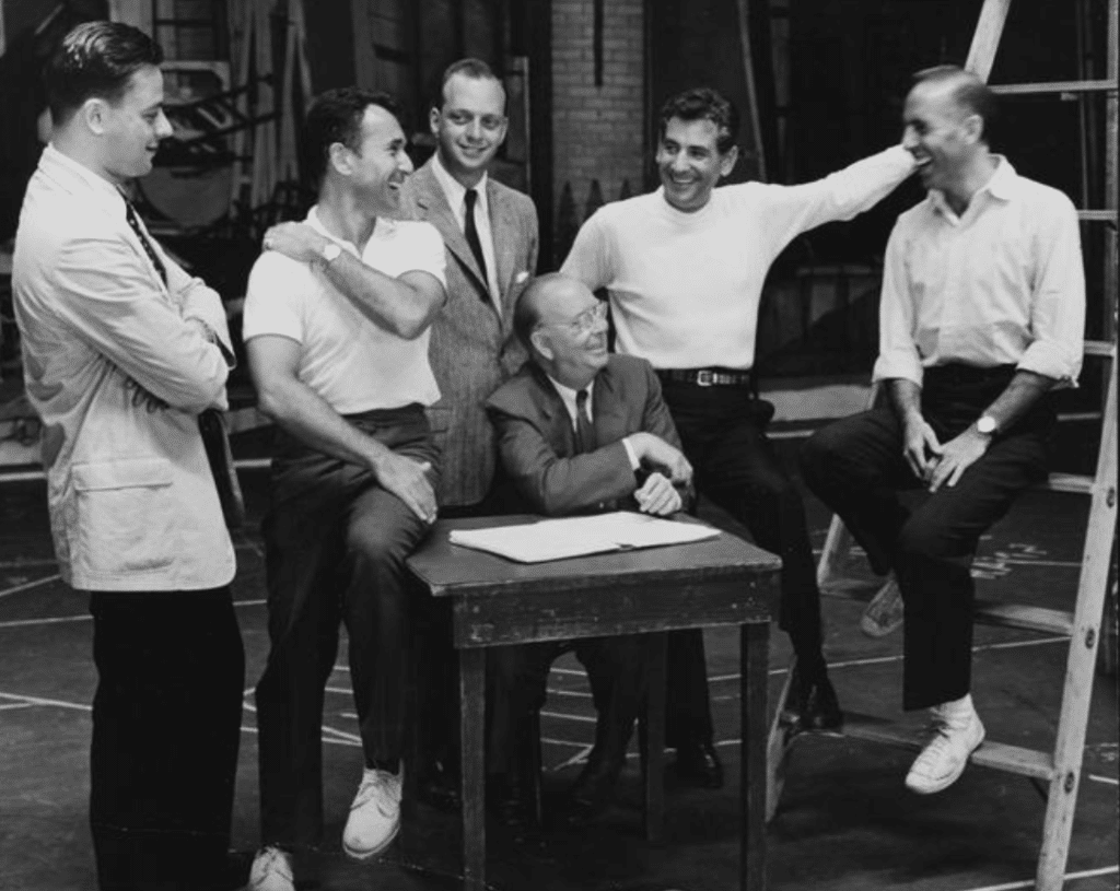 A man wearing a suit sits at a small table with an open book in front of him. Surrounding him are five other men wearing either suits or white shirts with black pants. One of the men is sitting on the table, another is sitting on a nearby ladder. They are all smiling and looking at each other. Black and white photo.