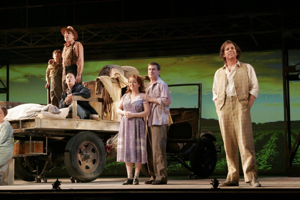 On a stage, six men and one woman wearing depression-era clothing stand on or near an old flatbed truck, some with their mouths open as if singing. The backdrop shows a green field and sky with yellow clouds.
