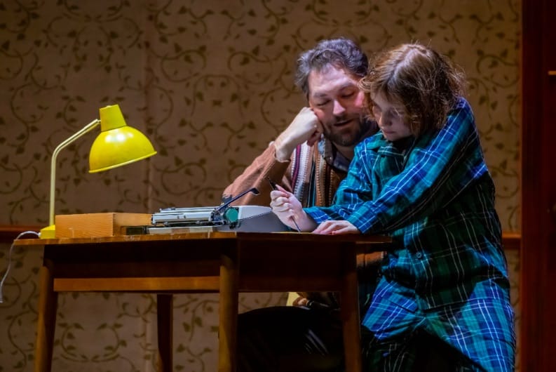 A child wearing green and blue pajamas sits on the knee of a man at a desk, both looking at something the child is writing with a pencil. On the desk are a yellow lamp and a typewriter.
