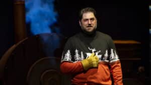 A man wearing a red and black sweater with white pine trees on it holds a hand wearing a yellow work glove to his abdomen. He is making a grimacing face, and some blue smoke can be seen in the darkness behind him.
