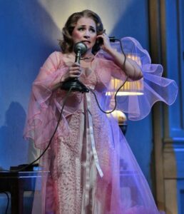 A woman in a long, pink dress with a sheer robe holds a candlestick telephone with a concerned look on her face.