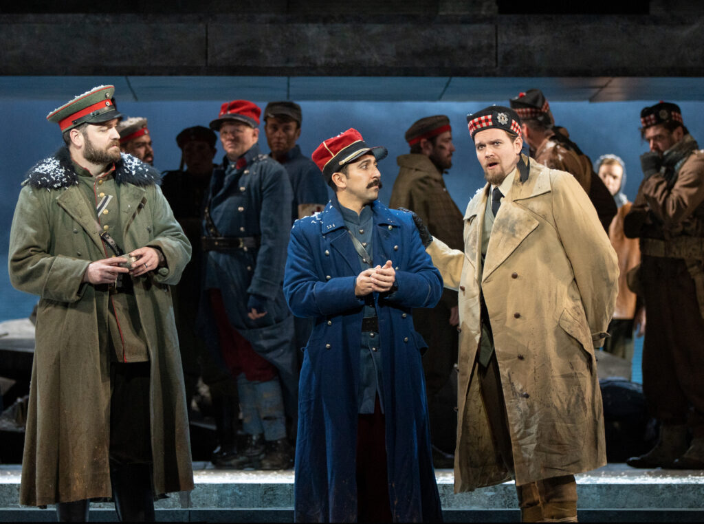 Several men wearing theatrical costumes in the style of early 1900s style military uniforms, hats, and jackets stand in a crowd. In the foreground, one man in tan is speaking to a man in blue while a third man in green looks on. Some have snow on their shoulders.