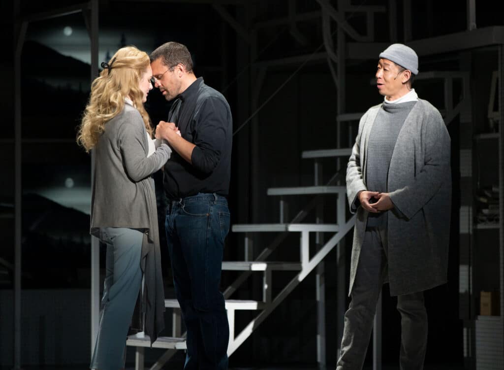 A woman and a man face each other holding hands with their foreheads touching. The woman has blond hair; the man is wearing a black turtleneck. Behind them, a man wearing all grey watches them with his mouth open as if singing.