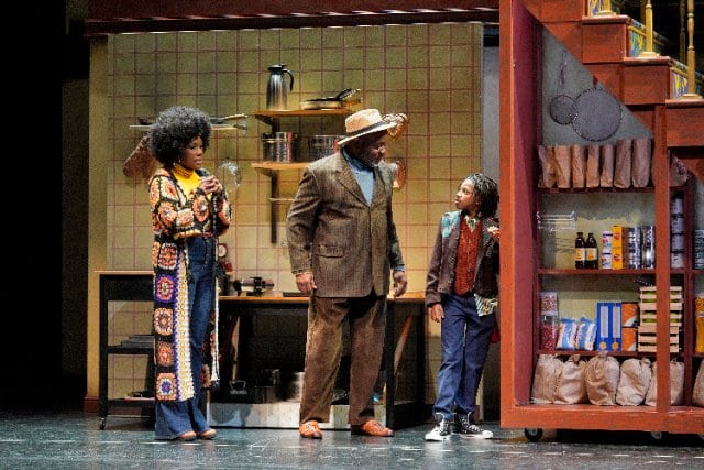 Three people stand in a yellow kitchen on a stage. A woman with a crocheted duster and a man in a brown suit both look at a child in jeans and a brown jacket.