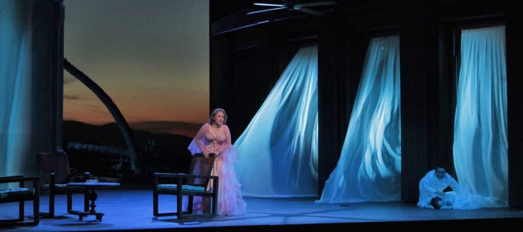 A woman in a long pink gown stands on a stage. To her right are chairs and a table, to her left are three long, white curtains that seem to be blowing in towards her. On the floor next to one of the curtains are two people in white: one lying on the ground and the other kneeling next to the first. The background is a sky with the last light of a sunset